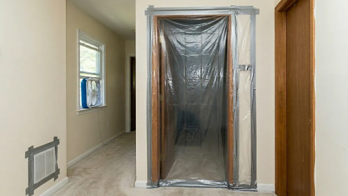 Mold remediation containment setup showing plastic sheeting taped over a doorway, a box fan in the window, and a sealed HVAC vent