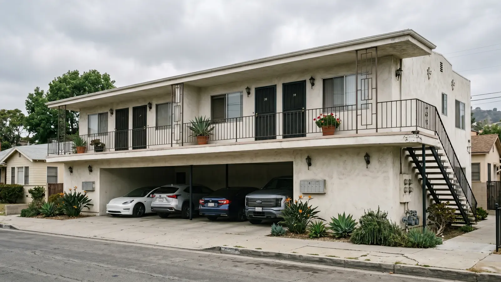 1960s dingbat-style stucco apartment building in Los Angeles showing carport parking on the ground floor, upper floor units with wrought iron railings, and exterior staircase