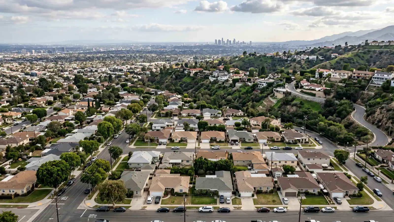 Aerial view of Los Angeles residential neighborhoods showing dense blocks of 1950s stucco ranch homes in the foreground, hillside properties with retaining walls mid-frame, and the downtown LA skyline visible in the hazy background