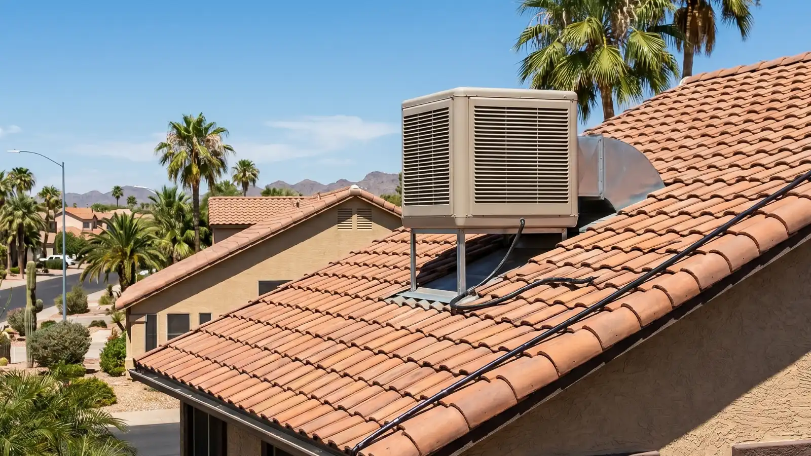 Evaporative cooler on a terra cotta tile roof in a Phoenix suburban neighborhood with saguaro cactus, palm trees, and mountains in the background