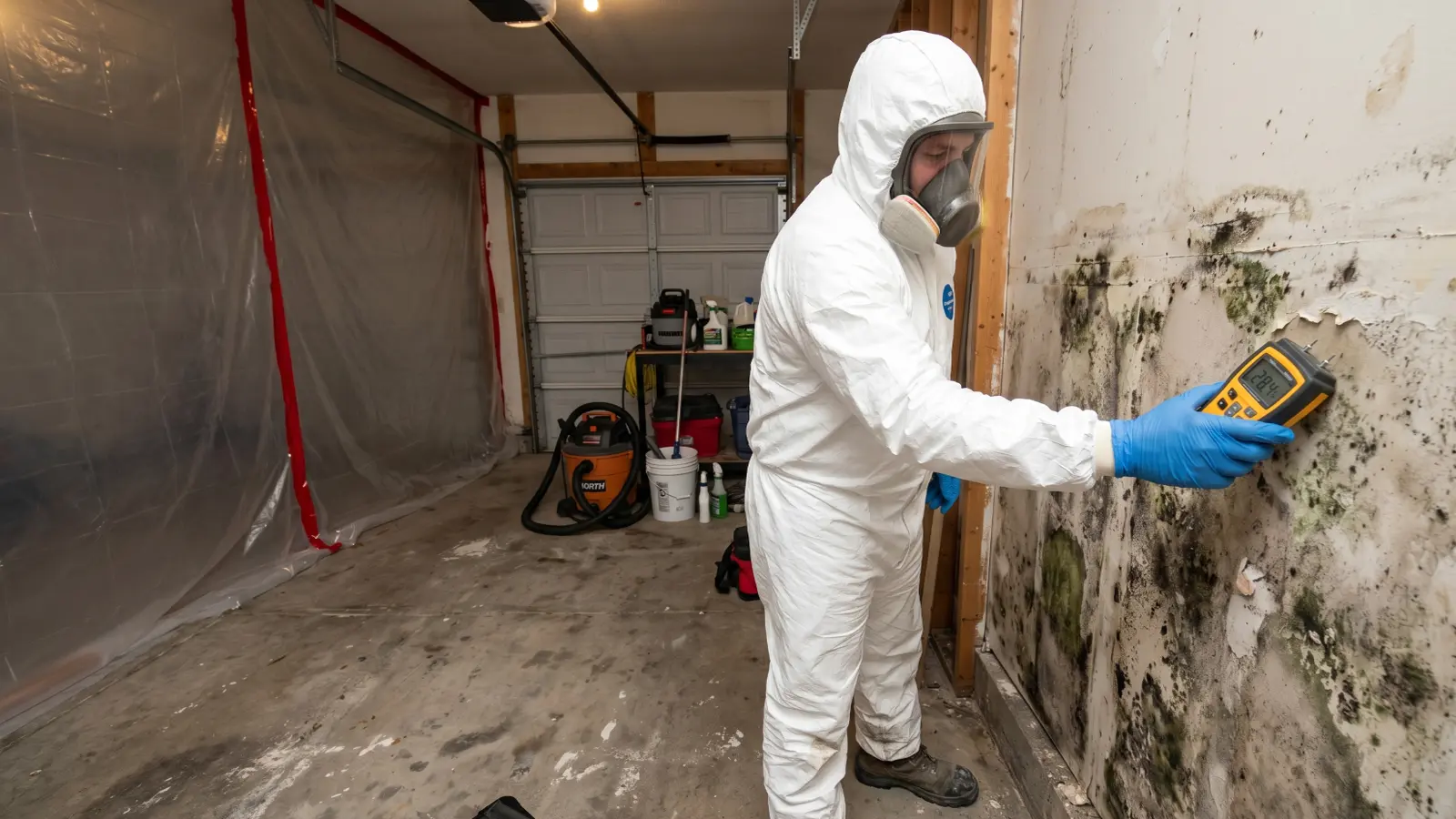 Professional mold remediation technician in Tyvek suit and respirator using a moisture meter on a mold-covered garage wall
