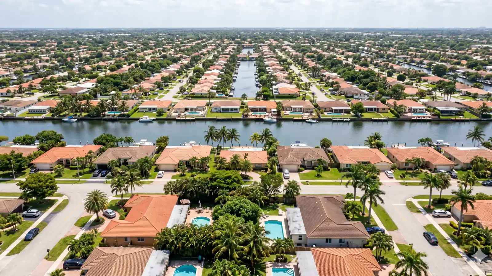 Aerial view of a Miami-Dade residential neighborhood with a canal running between rows of tile-roof homes, palm trees, and flat South Florida terrain stretching to the horizon