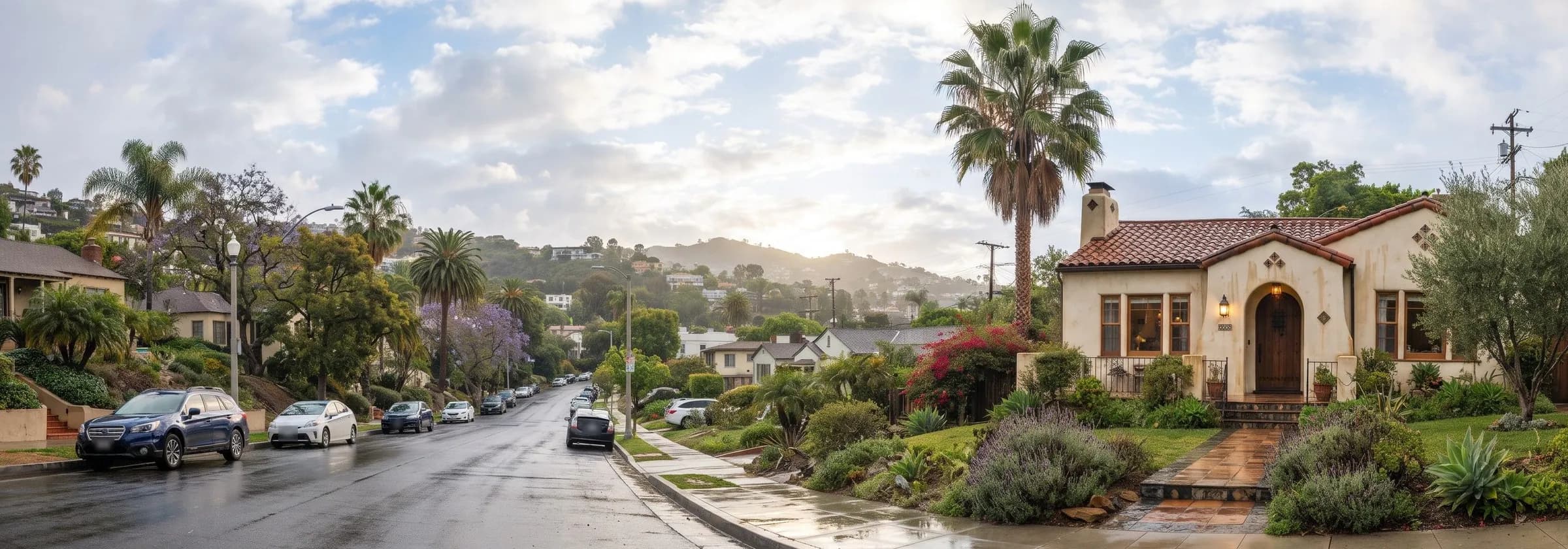 Single story stucco house with a red tile roof and an arched wooden front door beside a tall palm tree on a wet hillside Los Angeles street, with brown water stains running down the stucco below the roof edge