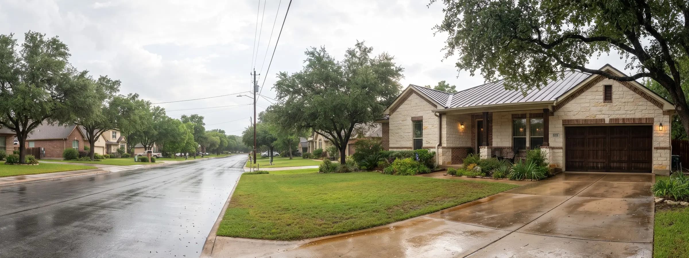 Single story limestone and brick house with a standing seam metal roof, a covered front porch with lanterns, and a two car garage with a dark wood door on a San Antonio street after rain, with a wet street, a damp driveway, and live oaks along the block